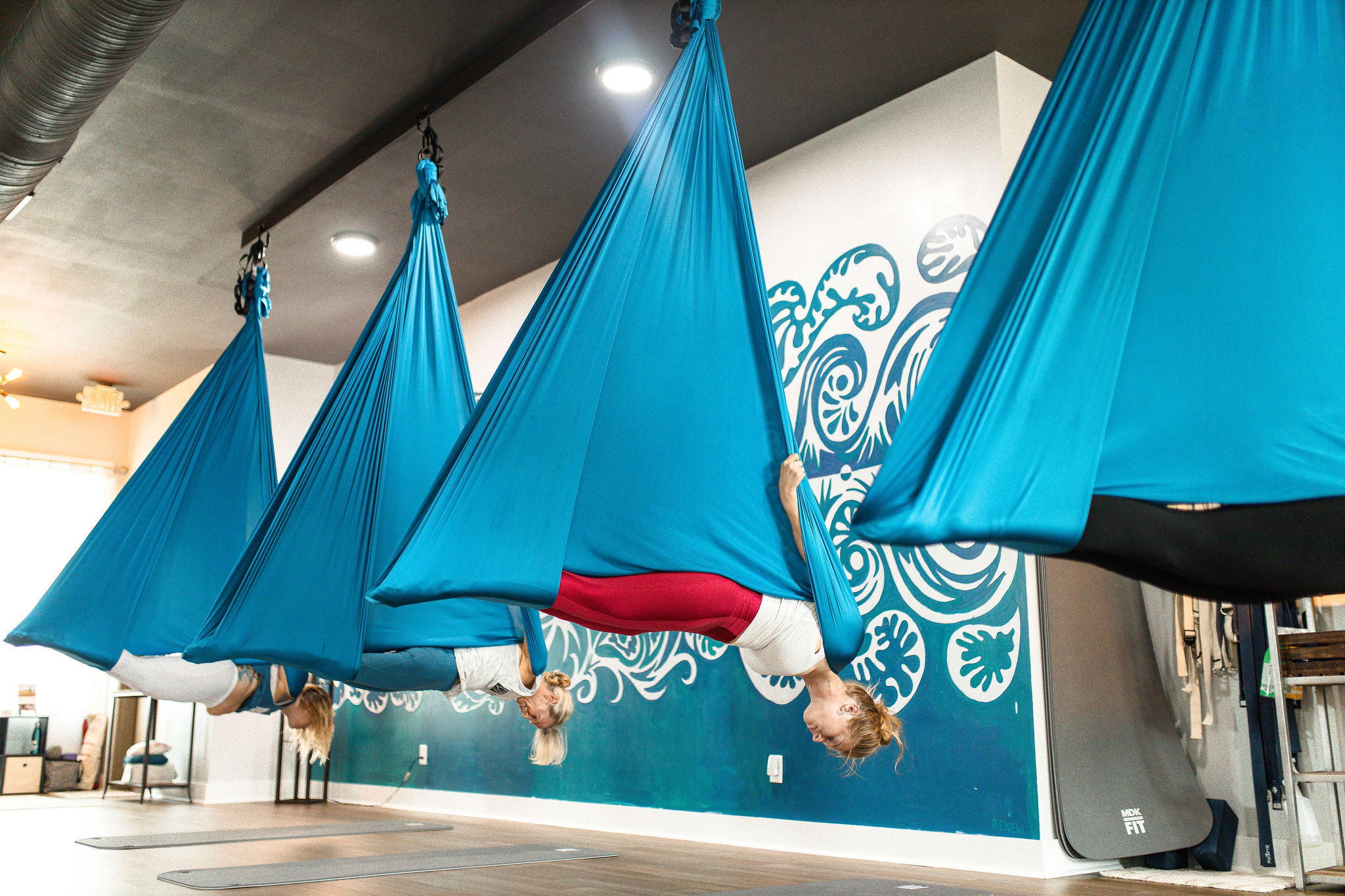 Group aerial yoga class with people suspended upside down in bright turquoise hammocks inside a modern indoor yoga studio with teal mural and mats on a wooden floor.