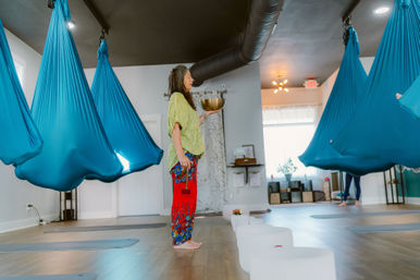 Bright aerial yoga studio interior with a barefoot woman holding a large singing bowl, turquoise suspended hammocks hanging from the ceiling, and a row of white crystal sound bowls on the wooden floor.