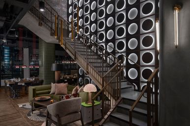 Modern industrial-chic hotel lobby interior with a brass staircase, dramatic backlit circular wall panels, cozy green sofa and armchairs, nearby dining tables with pendant lights, and warm accent lighting.