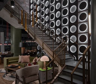 Modern industrial-chic hotel lobby interior with a brass staircase, dramatic backlit circular wall panels, cozy green sofa and armchairs, nearby dining tables with pendant lights, and warm accent lighting.