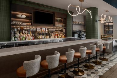 Sleek modern hotel bar interior with marble counter, row of leather-and-fabric swivel bar stools, backlit liquor shelves and taps, espresso station, green tiled columns, patterned floor tiles and sculptural pendant lights.