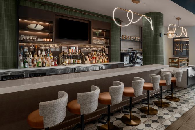 Sleek modern hotel bar interior with marble counter, row of leather-and-fabric swivel bar stools, backlit liquor shelves and taps, espresso station, green tiled columns, patterned floor tiles and sculptural pendant lights.