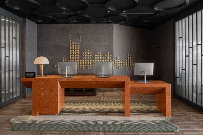 Contemporary boutique hotel reception in an urban lobby: terracotta check-in desk with three computer stations, brass lamp, gold geometric wall art, black circular ceiling accents and frosted glass partitions.