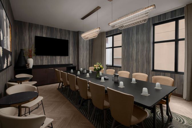Sleek modern conference room with a long black boardroom table surrounded by tan chairs, hanging tubular pendant lights, wall-mounted TV, wood-textured walls, two windows, sideboard with vase, small round café tables to the side, and a floral centerpiece with water pitchers on the table.