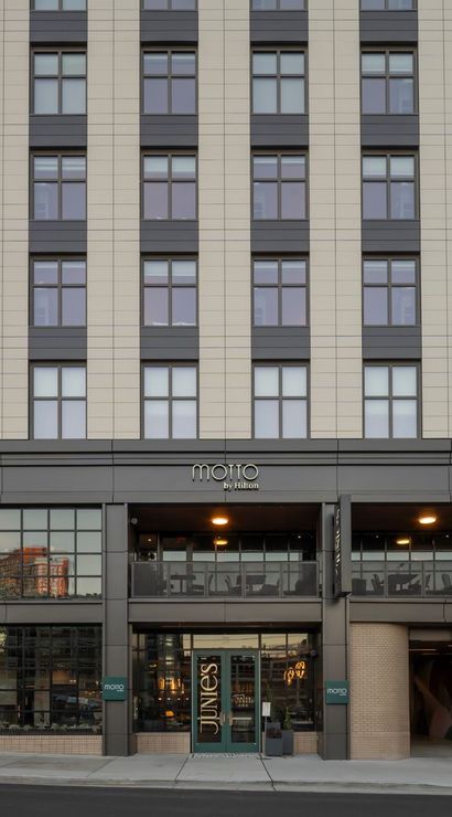 Sleek urban‑chic multi-story hotel facade with beige and gray cladding, repeating windows, ground-floor glass storefront and restaurant, balcony dining above the entrance, sidewalk and street in front.