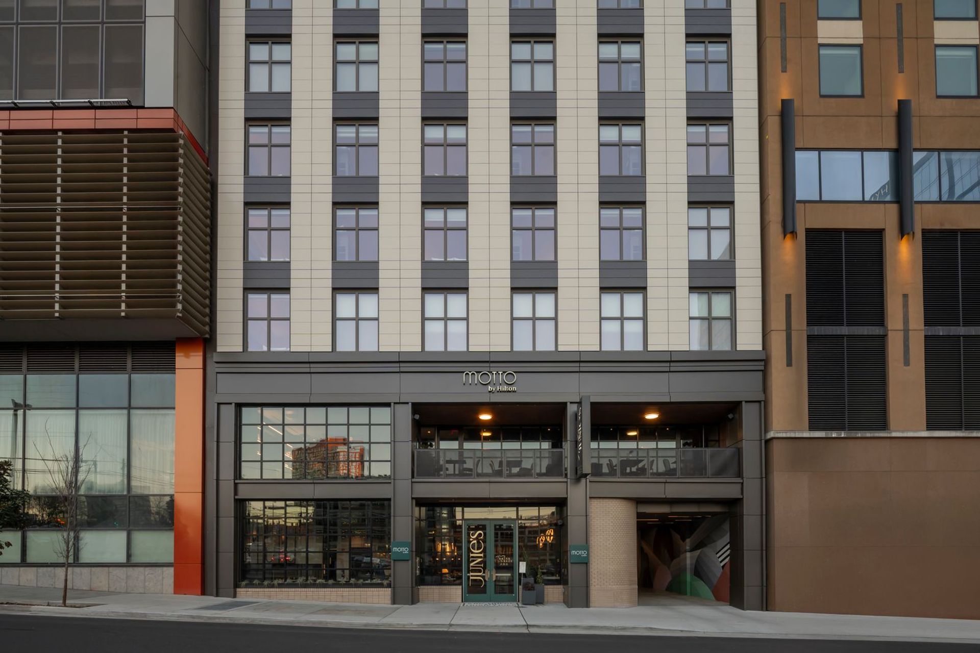 Sleek urban‑chic multi-story hotel facade with beige and gray cladding, repeating windows, ground-floor glass storefront and restaurant, balcony dining above the entrance, sidewalk and street in front.