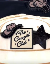 White cowgirl hat featuring a vintage-style script label, small embroidered hat patch and black glitter star embellishment.