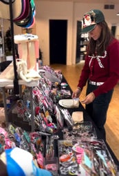 Woman in a green cap and red sweater browsing colorful hair accessories and jewelry on a boutique pop-up table inside an indoor shop