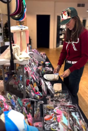 Woman in a green cap and red sweater browsing colorful hair accessories and jewelry on a boutique pop-up table inside an indoor shop