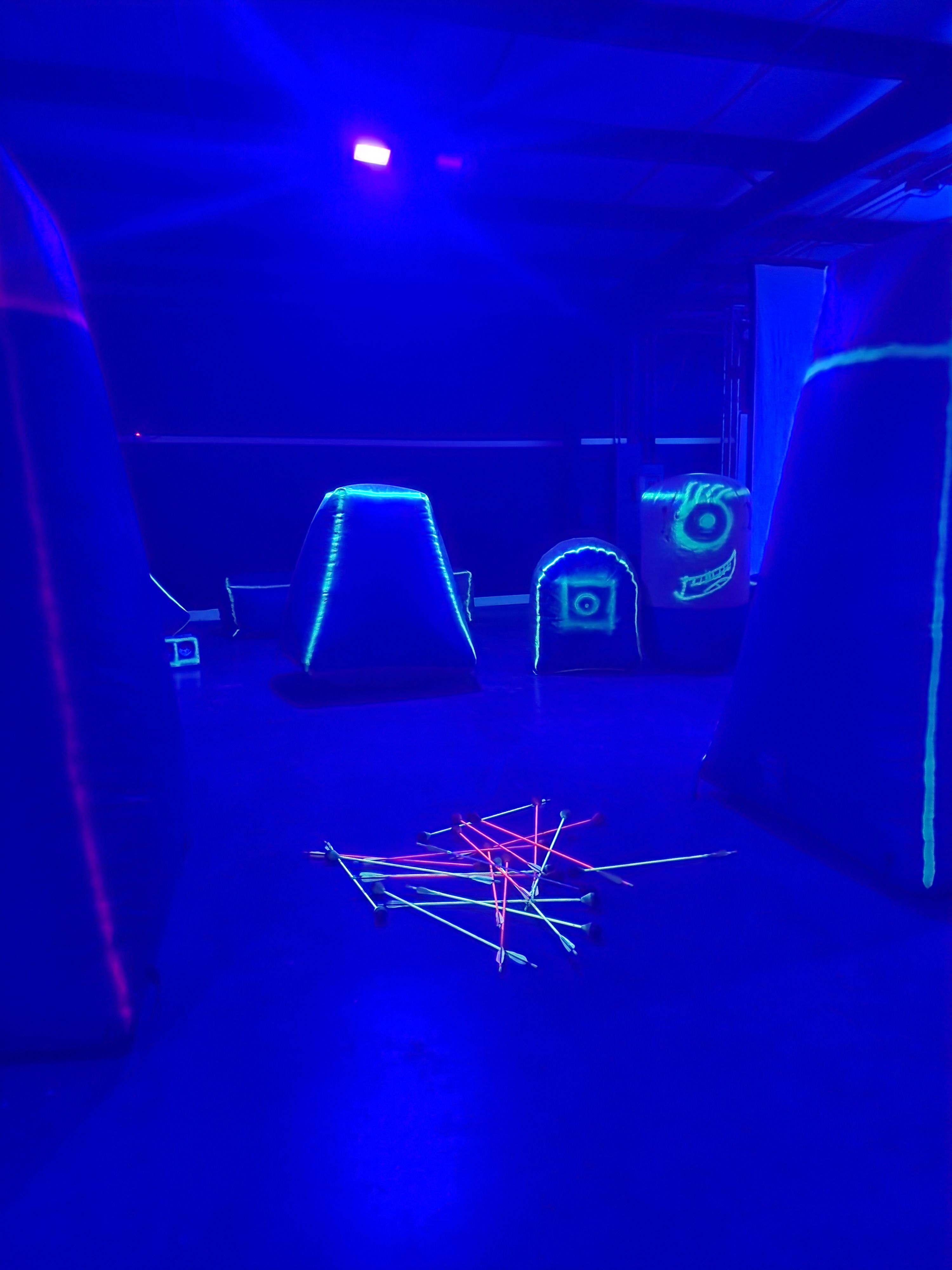 Neon blacklight indoor arena with inflatable bunkers and a scattered pile of glowing foam-tipped arrows on the floor under blue UV lighting