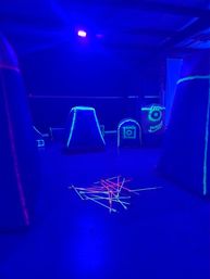 Neon blacklight indoor arena with inflatable bunkers and a scattered pile of glowing foam-tipped arrows on the floor under blue UV lighting
