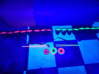 UV-lit neon archery gear: two bows, fluorescent arrows and three glowing target rings arranged on a black-and-white checkered floor