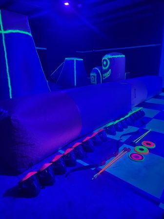 Indoor blacklight arena with neon inflatable bunkers and glowing bows, arrows, and target rings on a checkered floor, ready for glow archery fun