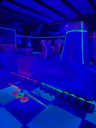UV-lit indoor arena with inflatable neon obstacles and glowing archery gear — bows, foam arrows and neon rings on a checkered floor