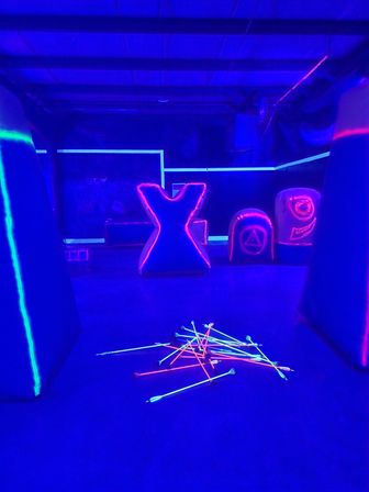 Neon-lit indoor archery arena under blacklight, glowing foam-tipped arrows scattered on the floor in front of pink-and-blue outlined inflatable bunkers and an X-shaped barrier.
