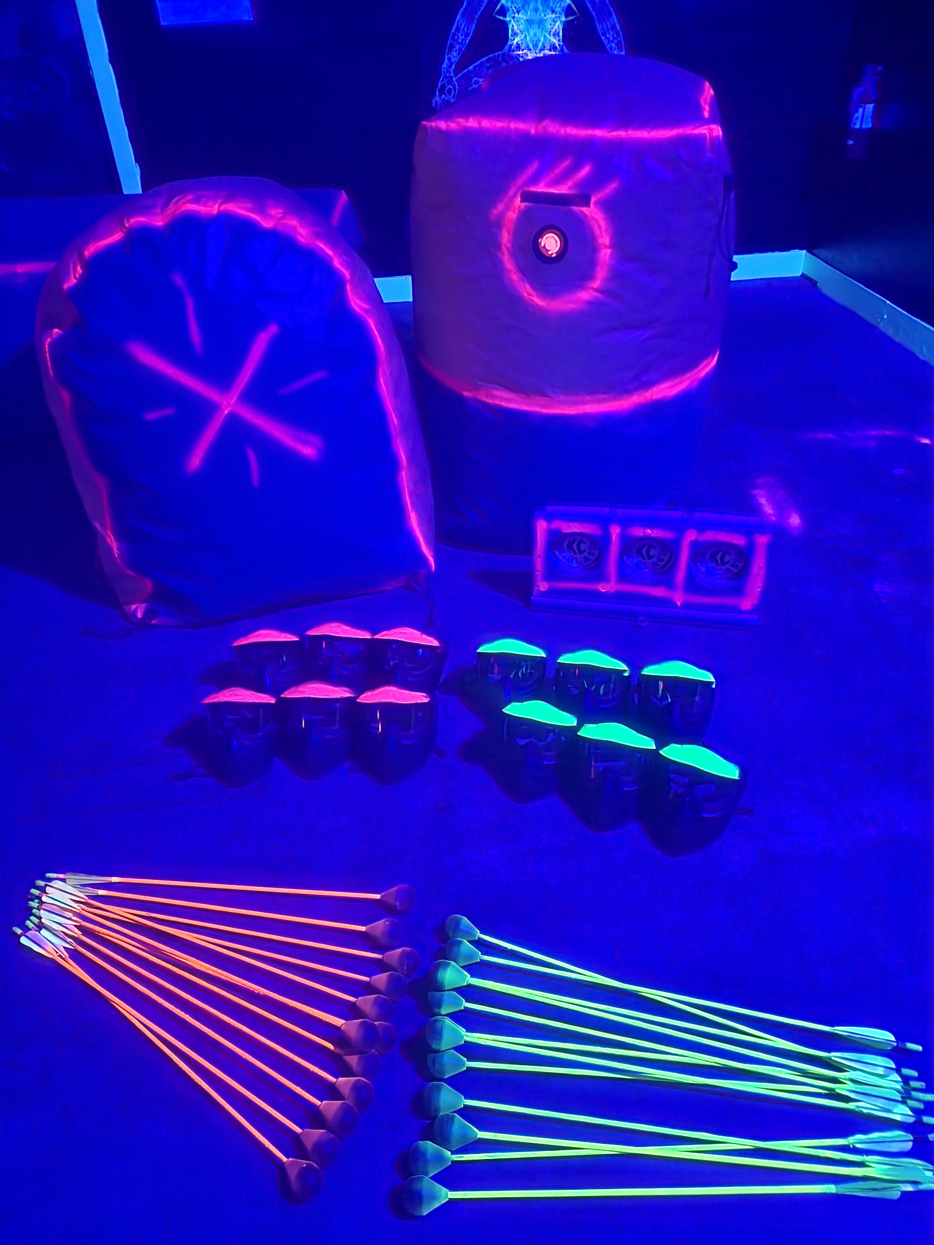 UV-lit indoor archery setup with rows of neon orange and green foam-tipped arrows and glowing padded bunkers and targets under blacklight, glow-in-the-dark equipment