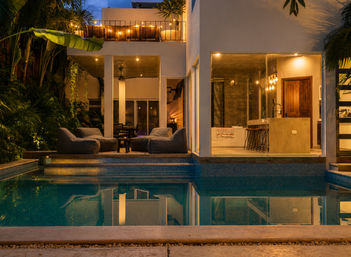 Evening view of a modern tropical villa with illuminated outdoor pool, cozy patio loungers, balcony string lights and an open-plan kitchen visible through floor-to-ceiling windows