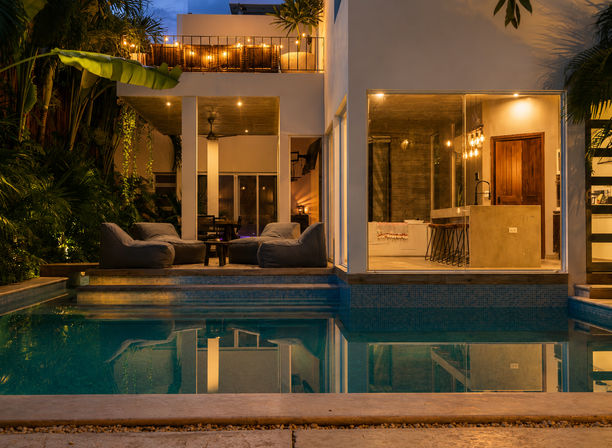 Evening view of a modern tropical villa with illuminated outdoor pool, cozy patio loungers, balcony string lights and an open-plan kitchen visible through floor-to-ceiling windows