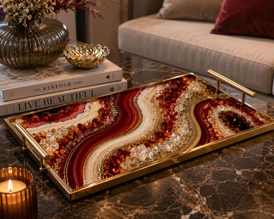 Eye-catching agate-style resin and crystal serving tray with red, white and gold swirls and brass handles on a marble coffee table, styled with stacked books, a glass vase of dried flowers, a gold bowl and a lit candle in a modern living room.