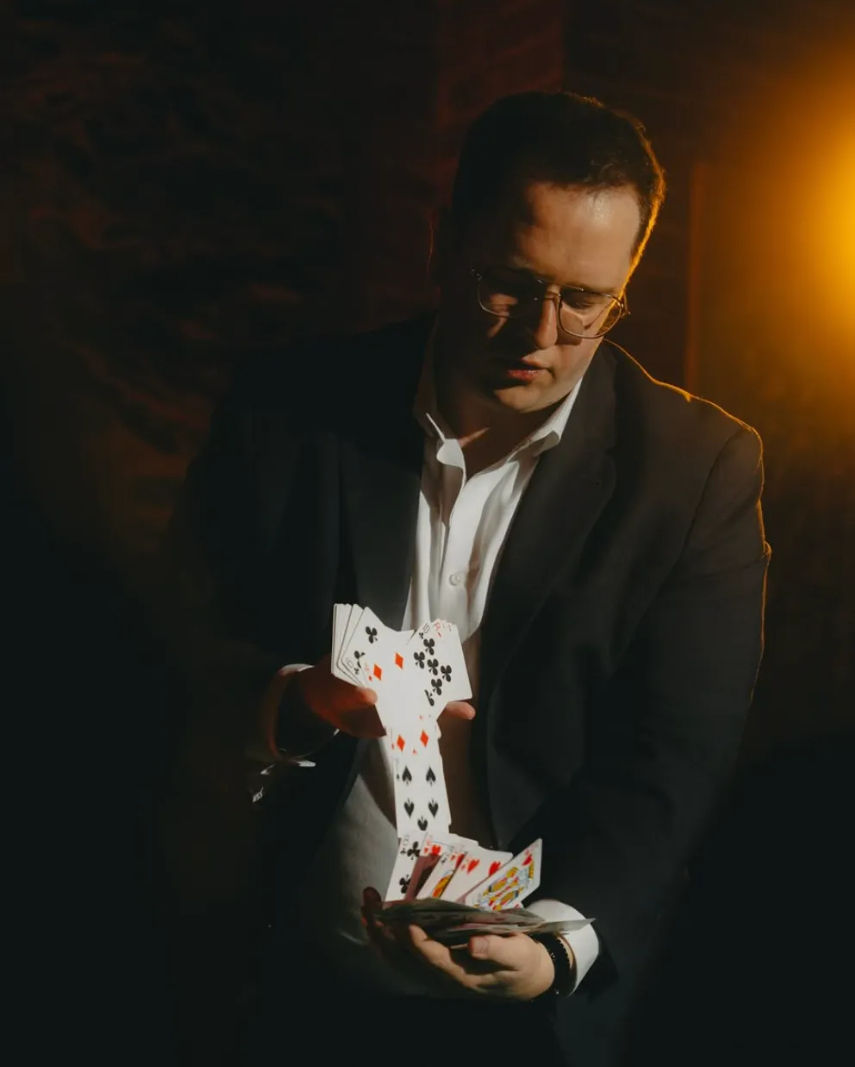 Person in a suit performing a sleight-of-hand card flourish, cascading playing cards in a dim, warm-lit indoor setting