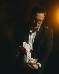 Person in a suit performing a sleight-of-hand card flourish, cascading playing cards in a dim, warm-lit indoor setting