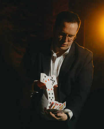 Person in a suit performing a sleight-of-hand card flourish, cascading playing cards in a dim, warm-lit indoor setting