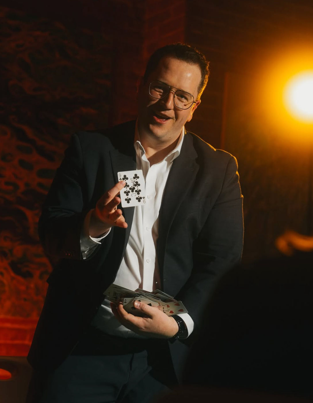 Magician in a suit and glasses performing a close-up card trick, holding a fan of cards and prominently showing the nine of clubs under warm stage lighting in an intimate indoor lounge.