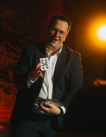 Magician in a suit and glasses performing a close-up card trick, holding a fan of cards and prominently showing the nine of clubs under warm stage lighting in an intimate indoor lounge.