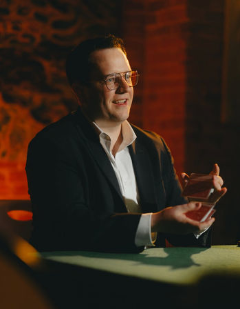 Smiling man in glasses and a suit performing a card trick with red playing cards at a green table in a warmly lit brick-walled room