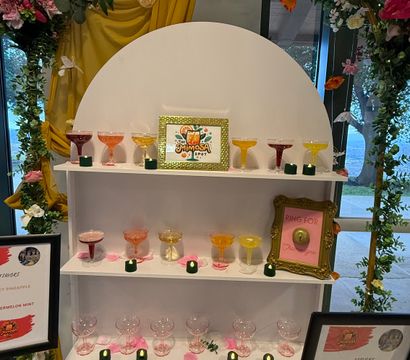 Indoor mimosa bar with a floral arch of white and pink roses and mustard-yellow drapery, tiered white shelves of colorful coupe glasses, scattered pink petals and small green votives.