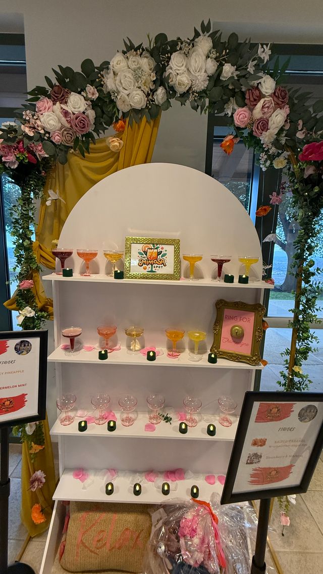 Indoor mimosa bar with a floral arch of white and pink roses and mustard-yellow drapery, tiered white shelves of colorful coupe glasses, scattered pink petals and small green votives.