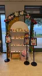 Indoor mimosa bar display with white arched shelving, colorful coupe glasses, framed menus, floral garland and yellow drape, scenic hillside visible through windows