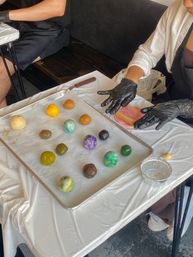 Colorful marbled dough balls arranged on a parchment-lined baking sheet while gloved hands roll pink dough on a plastic-covered table during a hands-on baking workshop