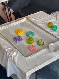 Vibrant pastel mooncakes arranged on a parchment-lined baking tray at a tabletop baking workshop, with colored dough balls on a white-covered table.