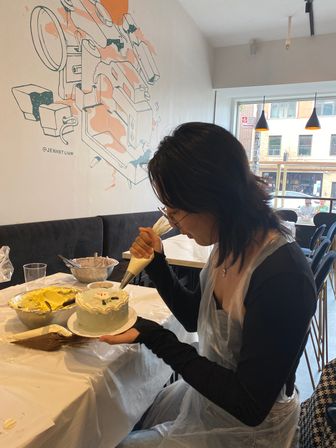 Participant piping frosting onto a small round cake during a hands-on cake-decorating workshop in a bright café, with bowls of buttercream and a muraled wall by large front windows.