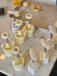 Cream-colored plastic cookie plunger cutters arranged on a marble kitchen counter with small dough balls and a parchment-lined baking tray, ready for home baking.