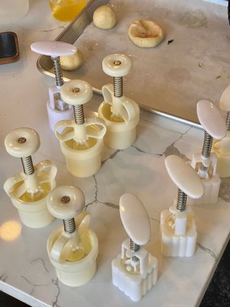 Cream-colored plastic cookie plunger cutters arranged on a marble kitchen counter with small dough balls and a parchment-lined baking tray, ready for home baking.