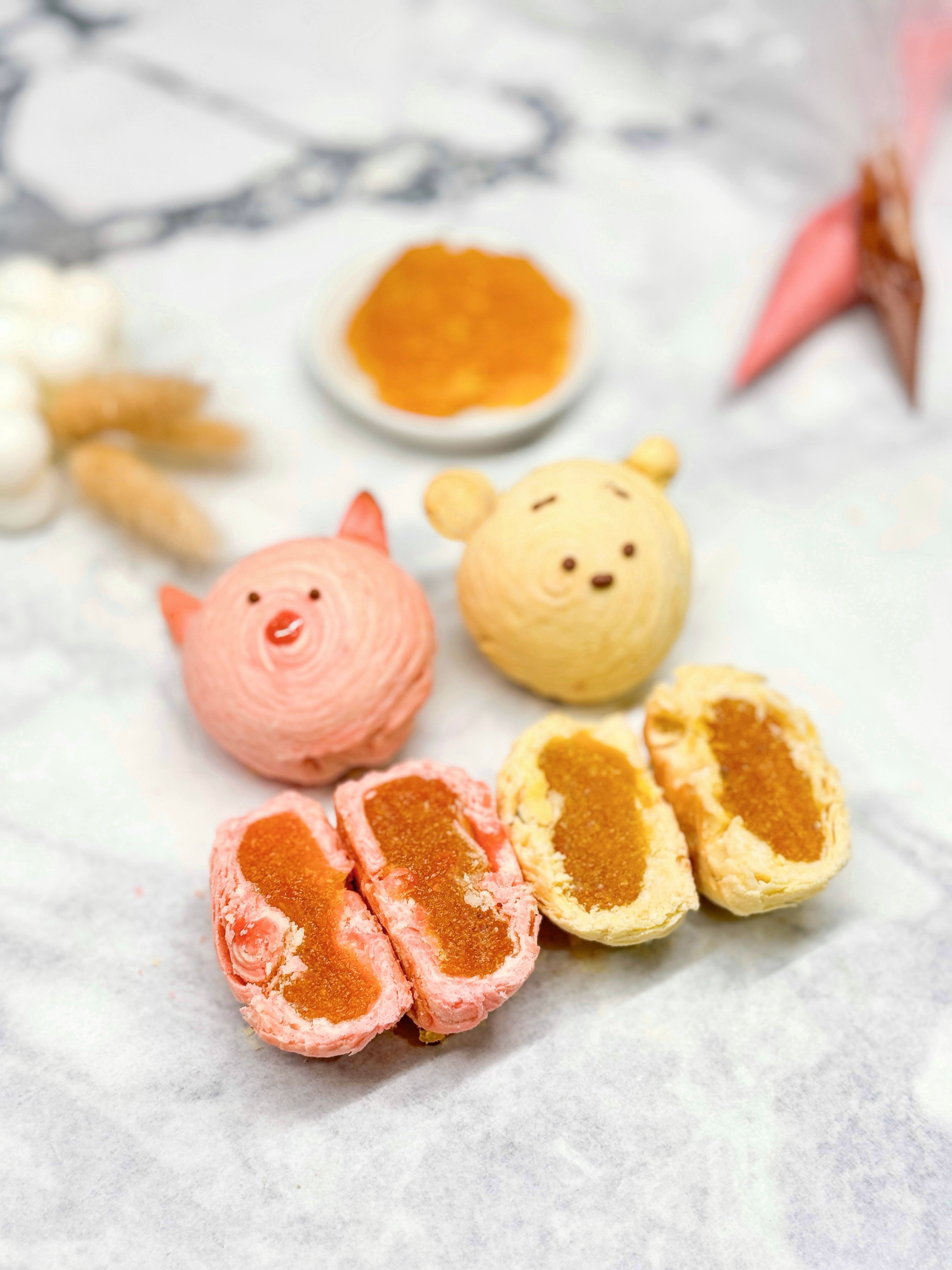 Close-up of cute pink pig and yellow bear swirl pastries with two halved flaky pineapple-jam-filled tarts on a marble surface — colorful Taiwanese-style pineapple pastries from a bakery.