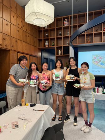 Six friends smiling in a wood-paneled craft studio, each holding a decorated dessert slice and plate after a fun cookie-decorating workshop, tables with icing supplies in foreground.