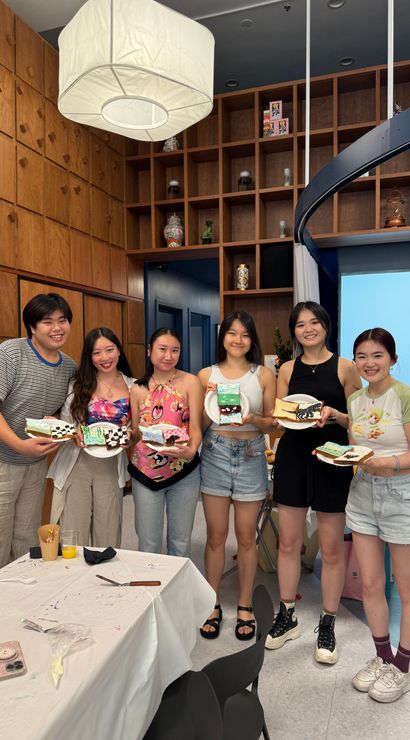 Six friends smiling in a wood-paneled craft studio, each holding a decorated dessert slice and plate after a fun cookie-decorating workshop, tables with icing supplies in foreground.