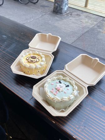 Two pastel mini character cakes in compostable takeout boxes on a dark wooden cafe table by a city sidewalk window