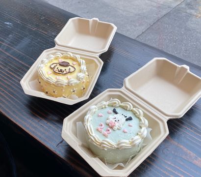Two pastel mini character cakes in compostable takeout boxes on a dark wooden cafe table by a city sidewalk window