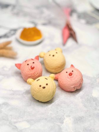 Four cute bear- and pig-shaped cream puff pastries in pastel yellow and pink on parchment over a marble countertop, small dish of orange dipping sauce blurred behind.