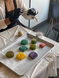 Artisan bakery scene: gloved hands pressing a mold to form colorful patterned mooncakes (green, teal, yellow, purple, pink) on a parchment-lined baking tray.