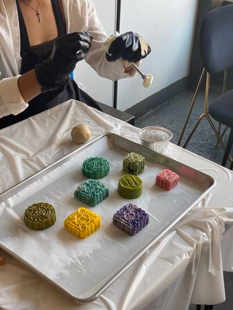 Artisan bakery scene: gloved hands pressing a mold to form colorful patterned mooncakes (green, teal, yellow, purple, pink) on a parchment-lined baking tray.