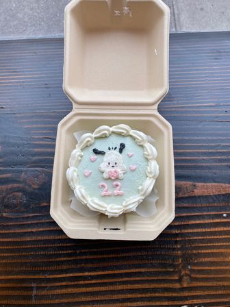 Mini pastel-green cake decorated with a cute animal face, pink hearts and the number "22", served in a beige takeout clamshell on a dark wooden table.
