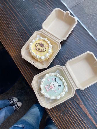 Two pastel mini cakes with cute animal-face icing (yellow and mint) in cardboard to-go boxes on a dark wooden table, whipped-cream borders and pink heart sprinkles — casual dessert takeout.