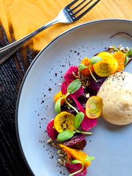 Vibrant farm-to-table beet salad with golden and red beet slices, pickled pink beet, microgreens, sunflower seeds and a creamy mousse dollop on a light gray plate with a fork on a wooden table