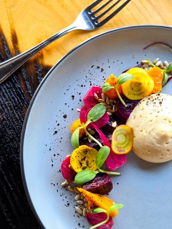 Vibrant farm-to-table beet salad with golden and red beet slices, pickled pink beet, microgreens, sunflower seeds and a creamy mousse dollop on a light gray plate with a fork on a wooden table