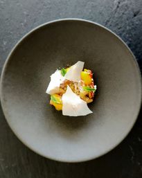 Top-down view of a fine-dining dessert in a matte black bowl: a small artful mound of shredded coconut and pastry topped with crisp white meringue shards, bright yellow fruit curd dollops and fresh mint leaves.